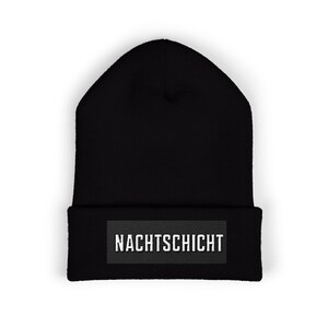 May include: Black beanie with a rectangular patch at the bottom. The patch has the word "NACHTSCHICHT" in white, sans-serif font. The beanie is made of a soft, knitted material.