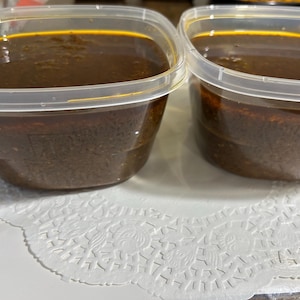 May include: Two clear plastic containers filled with a dark brown liquid. The containers have clear lids and are sitting on a white, patterned doily. The liquid appears to be a sauce or stew.