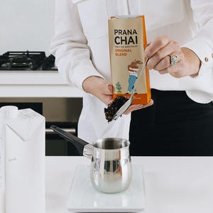 May include: A person pouring loose leaf tea from a Prana Chai bag into a stainless steel pot. The bag is orange and tan with the words "Prana Chai" and "Original Blend". A carton of milk sits to the left.