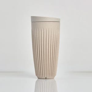 May include: A beige reusable travel cup with a speckled texture and vertical ribbed detailing along the exterior. The cup features a matching fitted lid and a tapered cylindrical shape, standing upright with a clean and simple design aesthetic.
