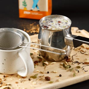 May include: A stainless steel milk frothing pitcher with a black handle, a white ceramic mug with a stainless steel tea strainer, and a wooden tray with tea leaves and spices. A box is in the background.