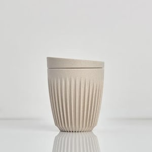 May include: A beige reusable cup with a speckled texture and vertical ribbed detailing along the exterior. The cup features a matching flat lid that sits securely on top, creating a clean and uniform appearance for this beverage container.