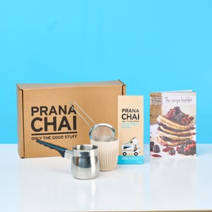 May include: A Prana Chai gift set featuring a brown cardboard box with black text, a stainless steel Turkish coffee pot with a black handle, a fine mesh strainer, a 250g bag of Peppermint Blend tea, and a recipe booklet with a pancake image.