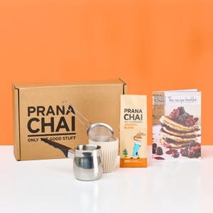May include: A Prana Chai tea set with a cardboard box, a bag of Original Blend tea, a recipe booklet, a stainless steel pot, a tea strainer, and a white cup. The box and tea bag have the words "Prana Chai" printed on them.