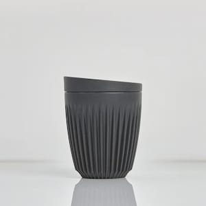May include: A matte charcoal gray reusable cup featuring a tapered silhouette with vertical ribbed texturing along the exterior body. The cup includes a matching dark gray lid that sits flush with the top rim, creating a clean and uniform look.