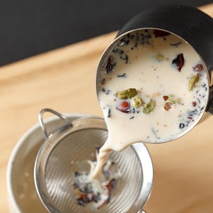 May include: A close-up of chai tea being poured from a small stainless steel pot through a mesh strainer into a white bowl. The chai tea is creamy with visible spices like cardamom and peppercorns.
