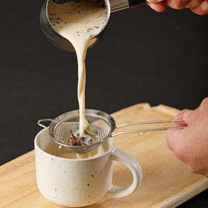May include: A stainless steel pouring pot with a black handle is pouring a creamy beige liquid through a fine mesh metal strainer into a speckled off-white ceramic mug. The strainer catches dark tea leaves as the liquid flows into the mug.