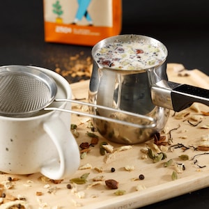 May include: A stainless steel Turkish coffee pot filled with a creamy beverage containing dark bits, paired with a fine mesh metal strainer resting on a speckled off-white ceramic mug. Loose tea leaves and spices are scattered across the wooden tray.