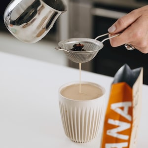 May include: A stainless steel pitcher pours tea through a mesh strainer into a beige ribbed ceramic mug. A bag with the word "PRANA" in white on an orange background is visible.