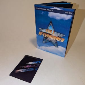 May include: A book titled "Full Afterburner Ace Combat Art" with a blue sky and cloud design. The cover features a star-shaped logo. A smaller card with a similar design is also present. The text "Patrick King Art Presents" and "Vol 003" are visible.