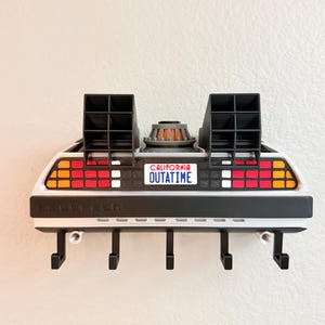 May include: A wall-mounted key holder shaped like the rear of a silver sports car. It features two black vents, a central circular engine detail, red and orange taillights, and a white license plate that reads CALIFORNIA OUTATIME. Five black hooks.