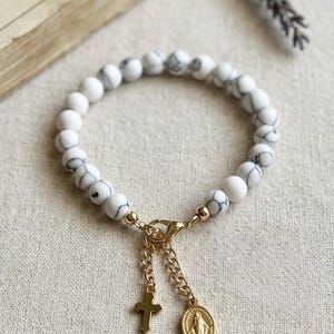 May include: A stretch bracelet composed of round white beads with grey veining. The bracelet features a gold-toned lobster clasp connecting two short gold chains, each holding a small gold charm: one a simple cross and the other an oval medal.