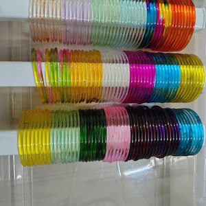 Op de afbeelding: Een verzameling kleurrijke glazen armbanden in een doorzichtige plastic container. De armbanden zijn in rijen gerangschikt en zijn er in verschillende kleuren, waaronder oranje, blauw, groen, roze en geel. De armbanden worden op witte steunen getoond.