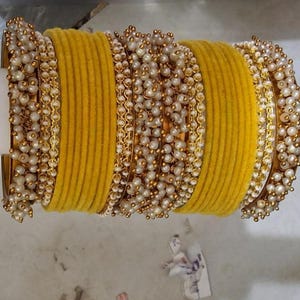 May include: A set of mustard yellow velvet-textured bangles arranged in a stack. The set features alternating bands of plain yellow velvet and gold-toned metal bangles densely encrusted with small, round, off-white faux pearls.