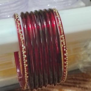 以下が含まれることがあります： 金色の宝石がちりばめられた縁取りのある、赤いガラスのバングルが積み重ねられています。バングルは白い表面に積み重ねられており、光がガラスの滑らかで光沢のある表面に反射しています。バングルは伝統的なタイプのジュエリーです。