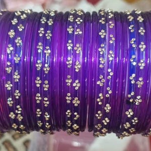 May include: A collection of purple bangles with gold-coloured floral embellishments. The bangles are made of a translucent material, and the gold accents are evenly spaced around each bangle. The bangles are stacked together, creating a visually appealing display.