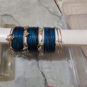 Op de afbeelding: Een set doorschijnende diepblauwe armbanden gerangschikt op een witte cilindrische houder. De collectie bevat verschillende effen blauwe glazen armbanden afgewisseld met dunne goudkleurige metalen armbanden met kleine bungelende bedeltjes.