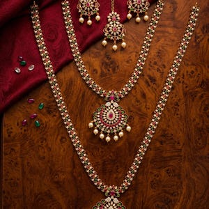 May include: A gold-toned jewelry set featuring two long necklaces with large circular pendants, a pair of matching earrings, and a maang tikka. Each piece is adorned with small white, red, and green stones and finished with dangling white pearls.