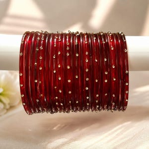 Puede incluir: Una pila de brazaletes de vidrio rojo oscuro, cada uno adornado con pequeños adornos brillantes. Los brazaletes se exhiben sobre un soporte cilíndrico blanco, mostrando su rico color y detalles intrincados. La joyería es un accesorio tradicional.