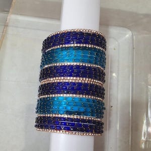 May include: Stack of blue and turquoise bangles with a decorative band of small, clear stones. The bangles are a mix of dark and light blue, with a band of turquoise in the middle. The bangles are stacked on a white cylinder.
