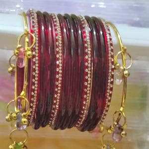 以下が含まれることがあります： 小さな透明なラインストーンが並んだ赤いガラスのバングル。金色の金属アクセントには、カラフルなビーズが付いた小さな揺れるチャームが含まれています。バングルは白い表面に積み重ねられています。