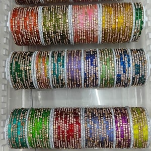 Op de afbeelding: Een verzameling veelkleurige armbanden gerangschikt in vier horizontale rijen op witte cilindrische houders. Elke armband heeft een metallic afwerking met ingebedde heldere steentjes en kleine goudkleurige rechthoekige accenten voor een levendig geheel.