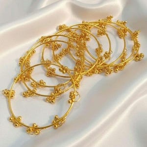 Op de afbeelding: Meerdere goudkleurige armbanden met een delicaat ontwerp van kleine, ronde kralen en bloemaccenten. De armbanden zijn gerangschikt op een witte, zijdeachtige stof, wat een luxueuze en elegante presentatie creëert. De sieraden zijn waarschijnlijk voor versiering.