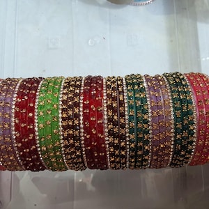 May include: A collection of colorful bangles in shades of pink, purple, red, green, and brown. Each bangle is adorned with small, sparkling embellishments, creating a dazzling display. The bangles are stacked together, showcasing their vibrant hues and intricate details.