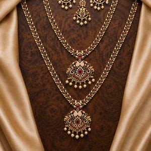 May include: A gold jewelry set featuring two long necklaces with intricate floral patterns, red and green stones, and white pearl accents. The set includes a pair of matching earrings and a maang tikka, all adorned with small white pearls.
