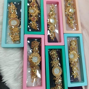 May include: Seven gold-toned wristwatches with intricate metalwork and colorful accents, each encased in a rectangular box with a clear window. The boxes are colored in shades of light blue, pink, teal, and peach, arranged in a cluster.
