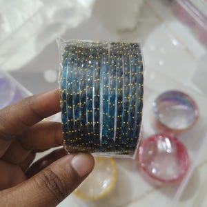 Puede incluir: Un conjunto de pulseras de cuentas azules y negras con detalles dorados. Las pulseras están envueltas en plástico transparente. Se ven otras pulseras coloridas en el fondo.