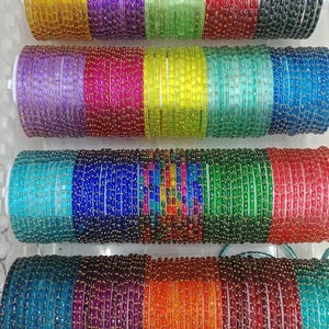 Op de afbeelding: Een verzameling dunne, ronde armbanden gerangschikt in vier horizontale rijen in een doorzichtige plastic bak. Elke armband heeft een getextureerd, doorschijnend oppervlak met kleine, gelijkmatig verdeelde goudkleurige accenten.