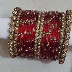 May include: A set of deep red translucent bangles featuring intricate gold-tone metal accents. The collection includes several thin red bands interspersed with wider, textured gold-tone bangles encrusted with small clear stones and white beads.