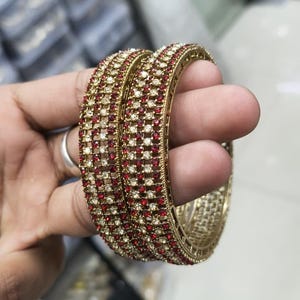 Peut inclure: Bracelets dorés avec des rangées alternées de pierres précieuses rouges et claires. Les bijoux sont tenus dans une main, mettant en valeur le design complexe et l'éclat des bracelets. Les bracelets sont un accessoire classique.