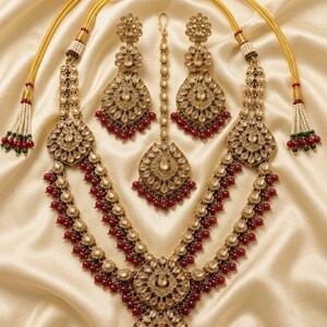 May include: A detailed gold-coloured jewellery set comprising a necklace, earrings, and a headpiece. The necklace has two layers with red bead accents. The earrings and headpiece echo the necklace design. The set is presented on a cream-coloured, satin-like fabric.