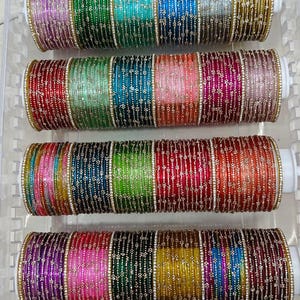 Op de afbeelding: Een verzameling veelkleurige armbanden gerangschikt in vier horizontale rijen in een doorzichtige plastic bak. Elke armband heeft een metallic afwerking met kleine, gelijkmatig verdeelde witte steentjes. De set bevat tinten paars, groen, blauw, zilver, goud, rood, oranje, roze en zwart.