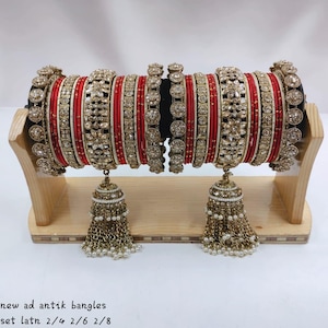 Pode incluir: Um conjunto de pulseiras vermelhas e douradas com padrões florais intrincados, pedras transparentes incrustadas e texturas metálicas. Dois grandes pingentes em forma de sino com contas pendentes estão nas laterais. Texto: new ad antik bangles set latn 2/4 2/6 2/8.