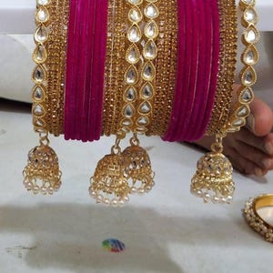 May include: A set of bangles featuring alternating bands of magenta velvet and gold-tone metal encrusted with small clear stones. The set includes two gold-tone metal bangles with teardrop-shaped clear stone accents and four hanging jhumka charms.