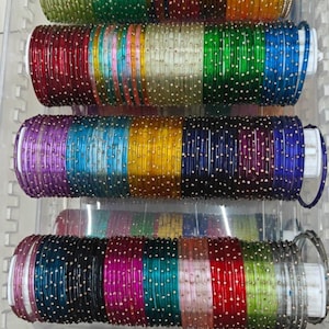 Op de afbeelding: Een verzameling dunne, ronde armbanden gerangschikt in vier horizontale rijen in een doorzichtige plastic bak. Elke armband heeft een gladde, doorschijnende afwerking in verschillende kleuren, waaronder groen, blauw, geel, rood, paars en roze, geaccentueerd met kleine, gelijkmatig verdeelde metalen stippen.