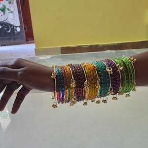May include: A selection of vibrant bangles in a range of colours, including red, orange, yellow, green, and purple, are displayed on a wrist. Each bangle is embellished with small, gold-toned charms. The bangles are stacked, creating a striking and colourful display.