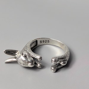 May include: A silver-colored ring with a rabbit design. The ring is open, with the rabbit's head on one end and its paws on the other. The ring is marked with "S925".