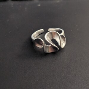 May include: A silver-colored ring with an open design. The ring features a flowing, abstract shape with curved lines and a polished finish. The ring is set against a dark background.