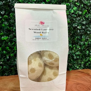 Peut inclure: Un sac en papier blanc avec une fenêtre transparente montrant plusieurs boules de laine blanc cassé avec des taches beiges. Le sac porte l'étiquette "Boho Beauty Scented Laundry Wool Balls" et "BABY SUDS".