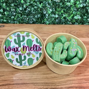 May include: A round container with a cactus pattern and the text "Wax Melts Savage Noir" next to a small paper cup filled with green cactus-shaped wax melts. The container lid is open, revealing the wax melts.