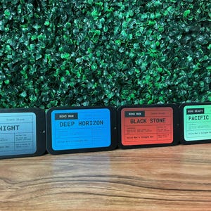 Puede incluir: Cuatro latas rectangulares de barras de colonia sólida. Las latas son negras con etiquetas en azul, rojo y verde. Las etiquetas dicen "Midnight", "Deep Horizon", "Black Stone" y "Pacific Drift". El fondo es una pared verde.