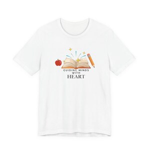 May include: White t-shirt featuring a colorful graphic. The design includes an open book, a red apple, and a pencil. The text "Guiding Minds with Heart" is printed below the book.