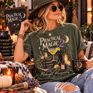 May include: A dark olive green t-shirt featuring a graphic print with the text Practical Magic 2 and Midnight Margaritas. The design includes a margarita glass, a black cat, stacked books labeled Salt, Rosemary, Lavender, Love, and a bottle labeled Spells.