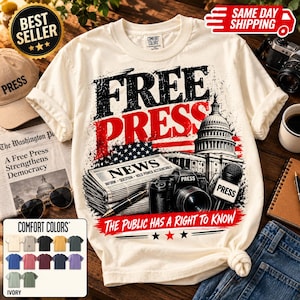 Puede incluir: Una camiseta de manga corta de color crema con un estampado gráfico con el texto FREE PRESS. El diseño incluye un periódico, una cámara, un micrófono con la etiqueta PRESS y el Capitolio de EE. UU., con el texto THE PUBLIC HAS A RIGHT TO KNOW.