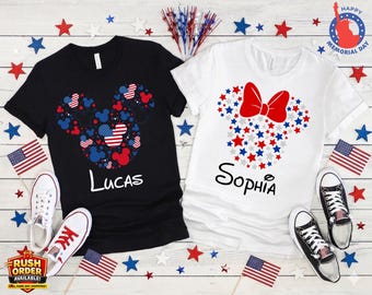 May include: Two t-shirts, one black and one white, with patriotic Mickey Mouse and Minnie Mouse designs. The black shirt features a Mickey Mouse head made of red, white, and blue shapes, with the name "Lucas" below. The white shirt has a Minnie Mouse head made of stars and a red bow, with the name "Sophia".