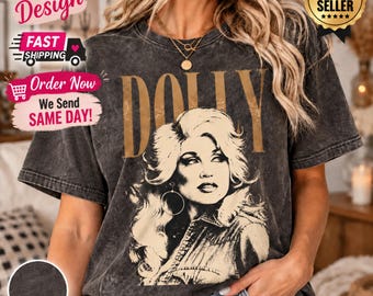 May include: A charcoal grey acid-wash t-shirt featuring a large, tan-coloured graphic print of a woman with voluminous hair and the word DOLLY printed in bold, tan capital letters above the portrait. The shirt has short sleeves and a crew neckline.
